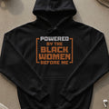 Black hoodie with "POWERED BY THE BLACK WOMEN BEFORE ME" text in orange and grey.